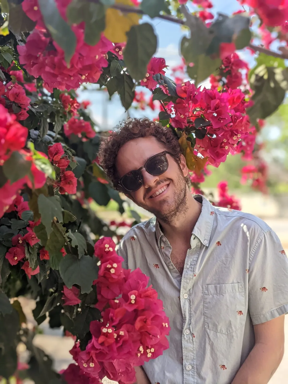 David grinning in sunglasses and a pale short-sleeved shirt, in front of a wall of bright pink bougainvillea.